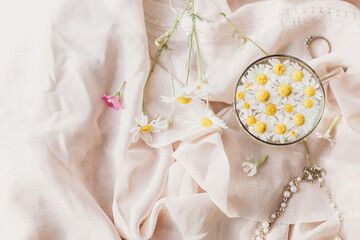 Obraz na płótnie Canvas Daisy flowers in water in glass cup on background of soft beige fabric with wildflowers and jewelry. Tender floral aesthetic. Creative summer image, flat lay. Bohemian mood