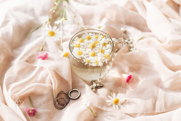 Daisy flowers in water in glass cup on background of soft beige fabric with wildflowers and jewelry. Tender floral aesthetic. Creative summer image with space for text. Bohemian mood