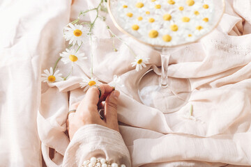 Hand with jewelry holding white flower on background of soft beige fabric with stylish wineglass with daisy flowers in water. Tender floral aesthetic. Creative summer image. Bohemian mood