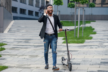 Smiling afro-american businessman with electric scooter walking near modern business centre and talking by phone. Modern technology and eco concept.