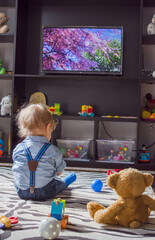 Cute baby boy and his teddy bear watching TV sitting on the floor covered with toys, at home