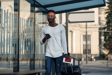 Young black male tourist with suitcase and passport in hands on bus of subway stop using internet maps in smartphone. Tourist finding a right way on smartphone in new city.