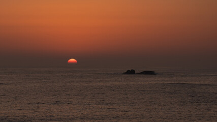 Sun disappearing below horizon at sunset on ocean with dark rocky islands in silhouette