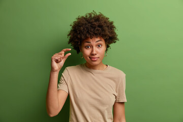 Fototapeta na wymiar Unimpressed curly woman shows some more or little bit gesture, demonstrates few inches or sentimeter, small amount or low scale sign, dresseed in casual clothes, isolated on green studio wall