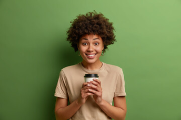 Glad curly haired Afro American woman drinks aromatic coffee from disposable cup, has interesting happy conversation, smiles toothily, wears casual clothes, isolated over vivid green background