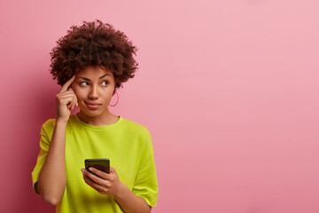 Pensive dark skinned woman keeps finger on temple, looks aside thoughtfully, uses new gadget for...