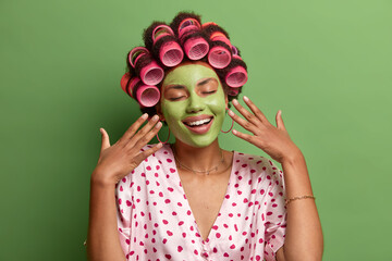 Pleased relaxed housewife stands with hands raised, closed eyes, gentle smile, applies moisturizing green mask on face hair rollers prepares for awesome event dressed in domestic clothes stands indoor
