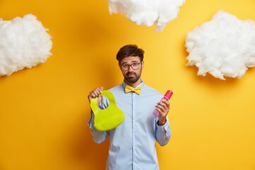 Photo of exhausted bearded father going to feed baby, holds bottle and bib, cares of newborn, dressed formally, poses against yellow background with white clouds. Fatherhood, parenthood concept