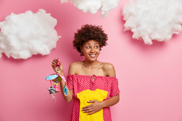 Happy ethnic pregnant woman holds yellow bodysuit and mobile for future baby, enjoys child...