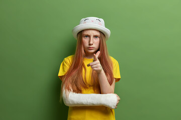 Small little girl with freckled skin and long ginger hair, points at you and looks seriously, wears hat and yellow t shirt, has broken arm after accidental fall, isolated on green background.