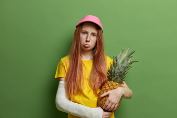 Sad offended freckled redhead girl being punished by parents, holds juicy pineapple, purses lips and looks gloomy at camera, got trauma during doing risky sport, poses against green background.