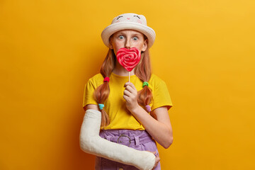 Surprised little girl covers mouth with big lollipop, enjoys summer time, has sweet tooth and holds delicious candy, dressed in stylish outfit, has broken arm, isolated on yellow background.