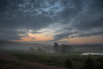 Foggy sunrise over a meadow and a river with dark clouds in the sky.