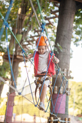 A little girl in an orange helmet and special protection goes through obstacles in a rope amusement...