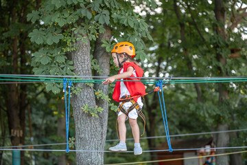 A little girl in an orange helmet and special protection goes through obstacles in a rope amusement...