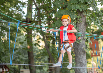 A little girl in an orange helmet and special protection goes through obstacles in a rope amusement...