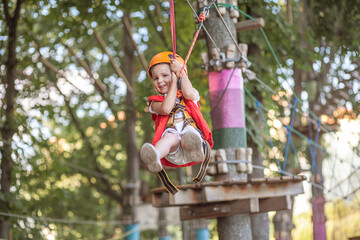 A little girl in an orange helmet and special protection goes through obstacles in a rope amusement...