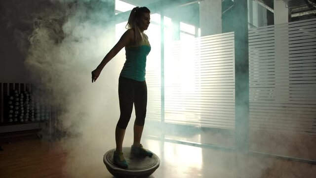 Caucasian attractive female is hardly squatting on the hemisphere in the gym. Hazed gym, large windows, silhouette of sexy woman body. Wide slow motion shot.