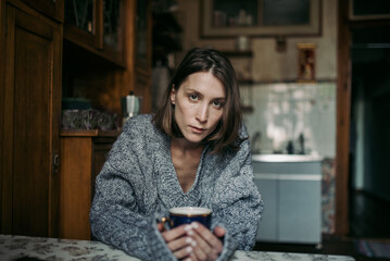 A young girl with a gray knitted sweater sits in the kitchen at the table with a cup in her hands