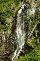 Obraz premium Top view of a beautiful waterfall on the mountain rock on a sunny summer day with a rainbow.