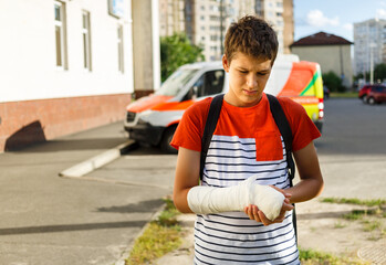 Teenage with broken hand in a cast in front of ambulance in the city. Boy in white t shirt holds his injured hand with arm splint.  