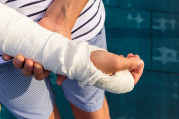 Cute Teenage with broken hand in a cast on sports ground in the city. Boy in white t shirt holds...