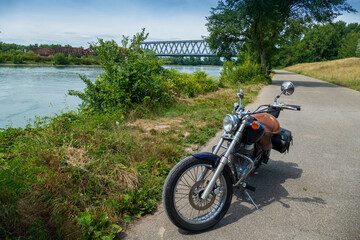 Fototapeta na wymiar Motorrad steht einsam am Rhein, Im Hintergrund die alte Brücke Wintersdorf