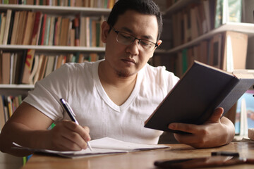 Young Asian man studying in library, exam preparation concept. Male college student doing research and making notes in his book