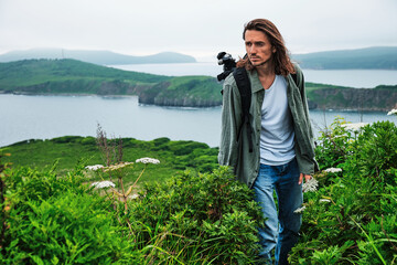 close up young long-haired man with a backpack and a photo tripod walking along a trail through the grass at the edge of a cliff with sky and sea background.. Travel and outdoor concept.