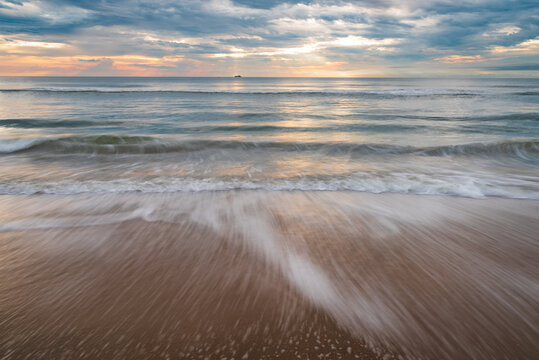 Sunrise on a serene beach. A long exposure shot of wave crashing on sand in Virginia Beach as the sun rises in a cloudy sky. 
