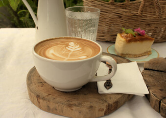 Latte coffee on a wooden serving tray with a picnic basket, glass of water and cheesecake in the background