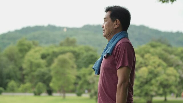 Asian middle age male take a rest, wiping sweat on his face and neck and drink water during exercise at the park, outdoor work out, body wellness tired exhausted, trees and mountain on the background