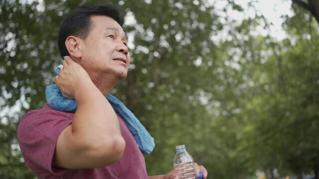 Thirsty Asian mature man take a rest while wiping sweat on his face and neck and drinking water during exercise at the park, standing under trees, body wellness tired exhausted, working out alone