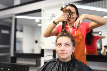 Woman make hair short. Client sit on chair in cape and watch hair cut. Cheerful hairdresser in apron hold bunch of hair for client and cut off with scissors. Do professional hair care and fashionable