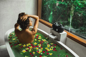 Beautiful young woman enjoying in spa, view from behind. Luxury stone bath tub with jungle view in window. Tropical flowers in water. Beauty treatment concept.