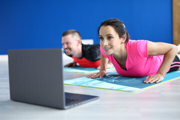 Couple look into laptop on floor. Woman and man do push-up on gymnastic mat and watch video in loptop. Drawing up a program for sport at home. Remote exercises online.