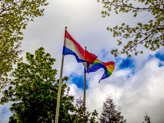 LGBT and Holland flags on poles blow in the wind. Symbol of tolerant. Gay sign rainbow. The rainbow pride, Trees and blue cloudy sky on the background. Copy space