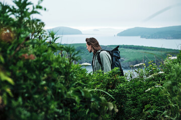 close up young long-haired man with a backpack and a photo tripod walking along a trail through the grass at the edge of a cliff with sky and sea background.. Travel and outdoor concept.