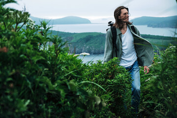 close up young long-haired man with a backpack and a photo tripod walking along a trail through the grass at the edge of a cliff with sky and sea background.. Travel and outdoor concept.