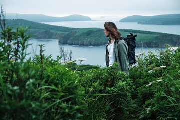 close up young long-haired man with a backpack and a photo tripod walking along a trail through the grass at the edge of a cliff with sky and sea background.. Travel and outdoor concept.