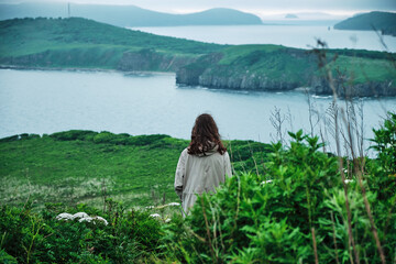 back view of a girl standing in the grass at the edge of a cliff with sky and sea background. Journey and outdoor concept.