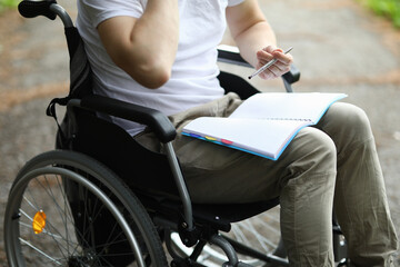 Obraz premium Man sit in wheelchair and take note in notebook in park. Disabled person keep diary and hold pen in his hand and notebook on his lap. Preparation of classes for rehabilitation after surgery.