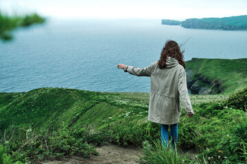 back view of a girl walking along a trail through the grass at the edge of a cliff with sky and sea background. Travel and outdoor concept.
