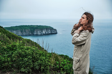 side view of a girl standing in the grass at the edge of a cliff with sky and sea background. Journey and outdoor concept.