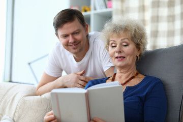 Mother sit on couch, hold photo album and show her son photo. Man relax and stand with his elbows on sofa behind woman. Elderly woman read book and show her son. Memories of childhood in photographs