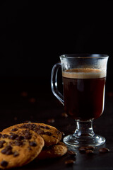 Close up of a glass cup of coffee, chocolate chip cookies and roasted coffee beans on dark background. Concept of ready to eat food, tasty snack. Selective focus, copy space.