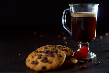 Close up of a glass cup of coffee, chocolate chip cookies and roasted coffee beans on dark background. Concept of ready to eat food, tasty snack. Selective focus, copy space.