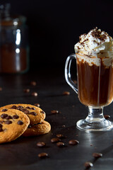 Close up of a glass cup of coffee with whipped cream and chocolate on it, chocolate chip cookies and roasted coffee beans on dark background. Concept of ready to eat food, tasty snack. Selective focus