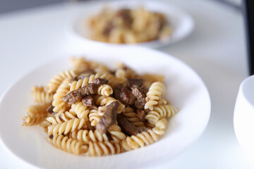 Pasta and meat are on plate. Two white plate of food stand on white table. Cooking delicacies at home. Food order online concept.