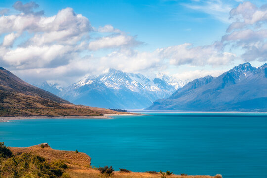 Spectacular view of snow-capped Mount Cook engulfed in foggy clouds with the blue waters of Lake Pukaki in the foreground on a beautiful summer day in Mount Cook NP in New Zealand's South Island.
